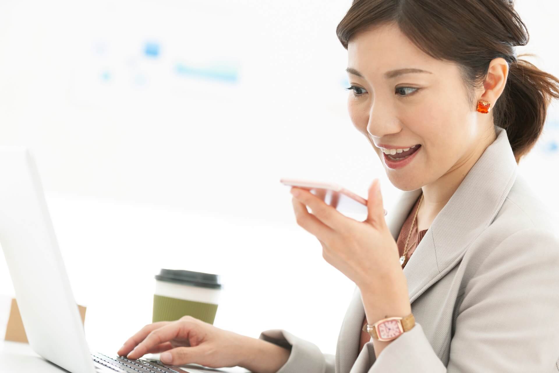 ノートパソコンの前でスマートフォンに向かって話しかけるビジネスパーソン