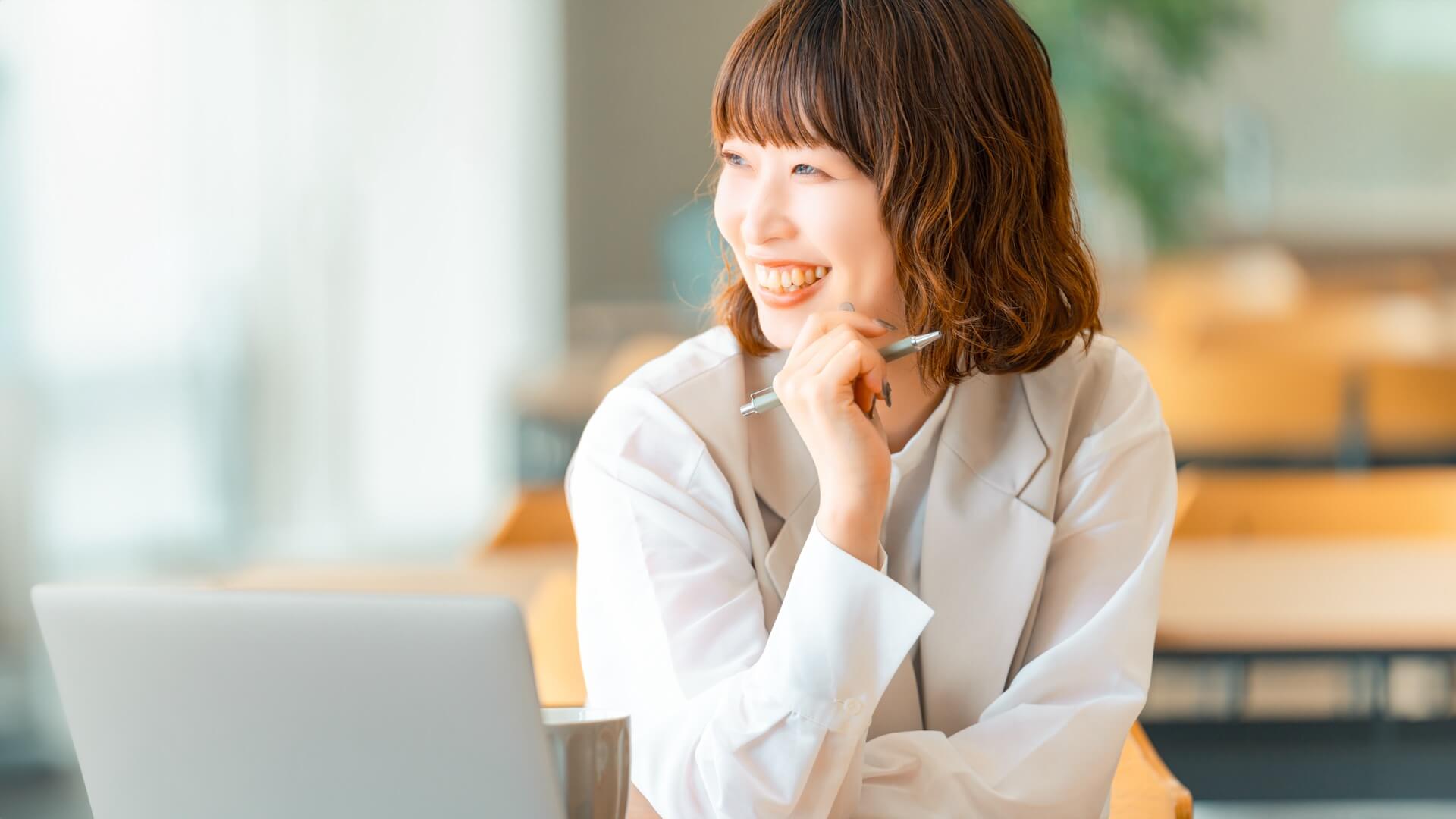 ノートパソコンの前でペンを持ち、笑顔で考えごとをする女性