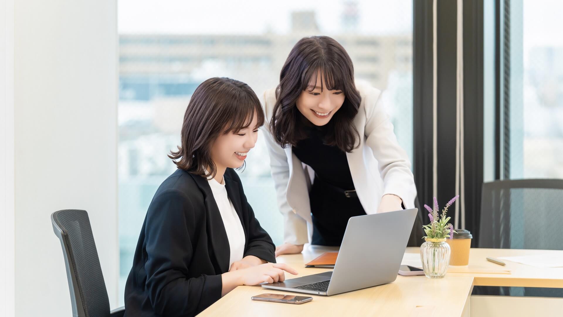 オフィスで1台のノートパソコンを2人でのぞき込み、笑顔で作業を進める女性たち