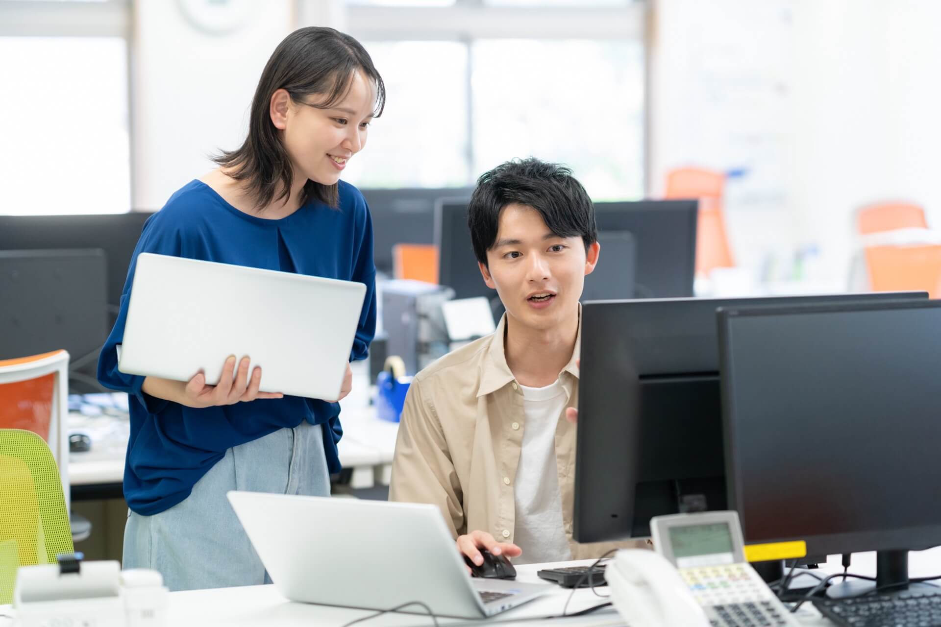 オフィスでノートパソコンを持った女性と、モニターに向かう男性が画面を見ながら話している様子