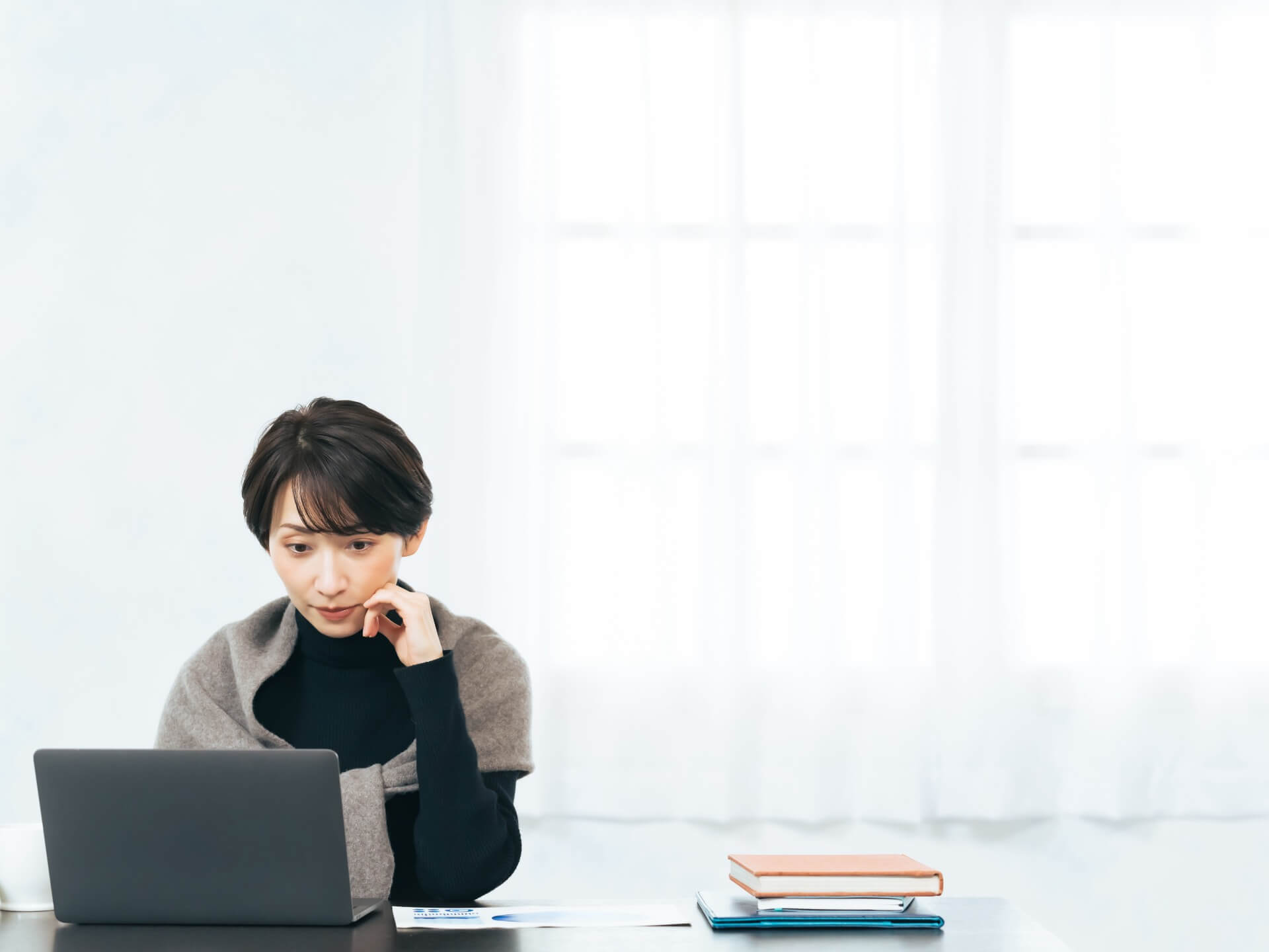 明るい部屋でノートパソコンに向かい、頬に手を当てて考えごとをする女性