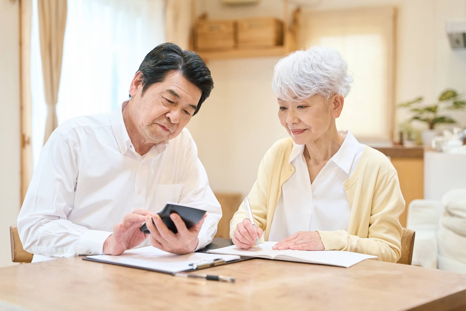 自宅のテーブルで、電卓を持つ男性とノートに書き込む女性のシニア夫婦が家計を計算している様子