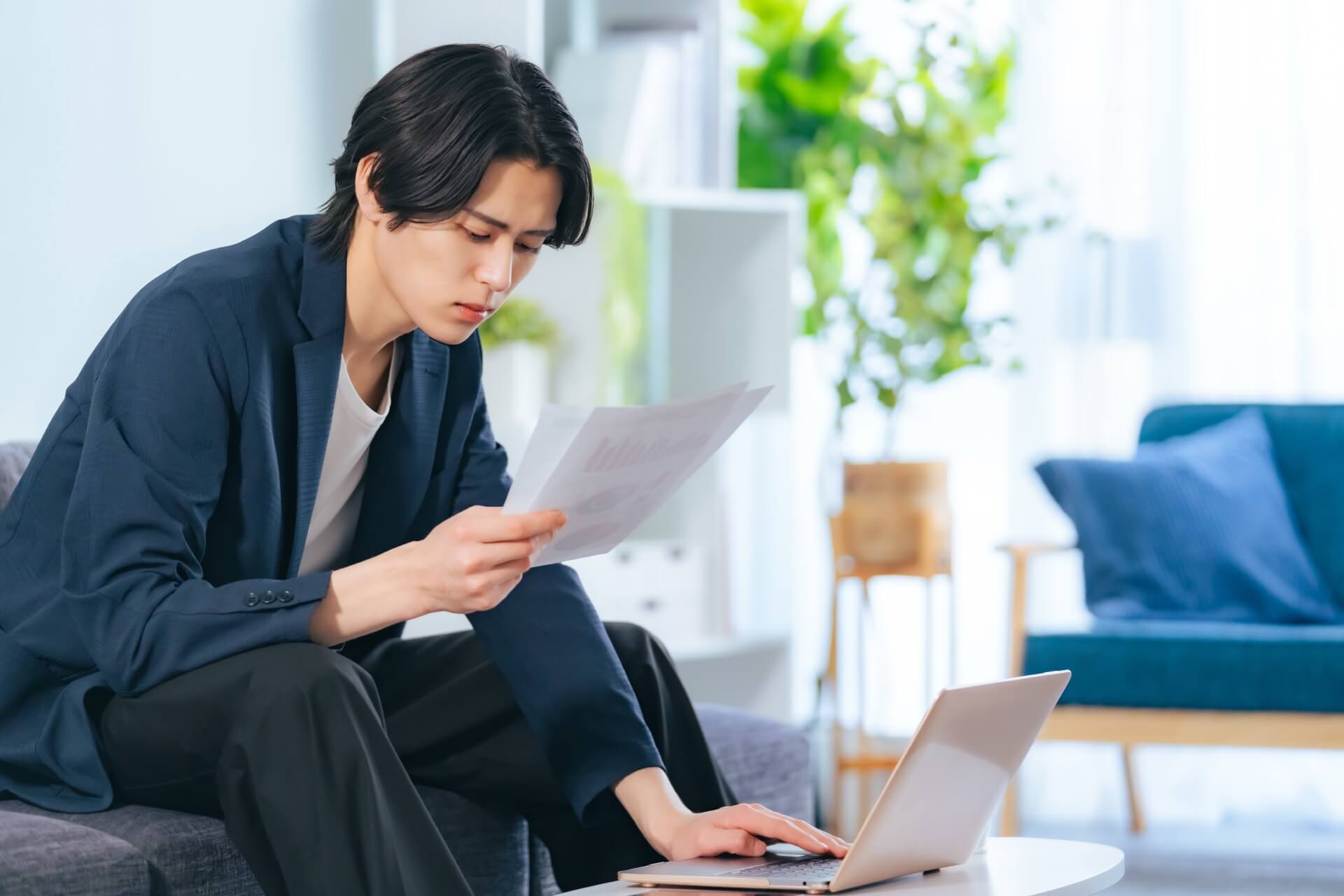 ノートパソコンの前で書類を読みながら考える男性