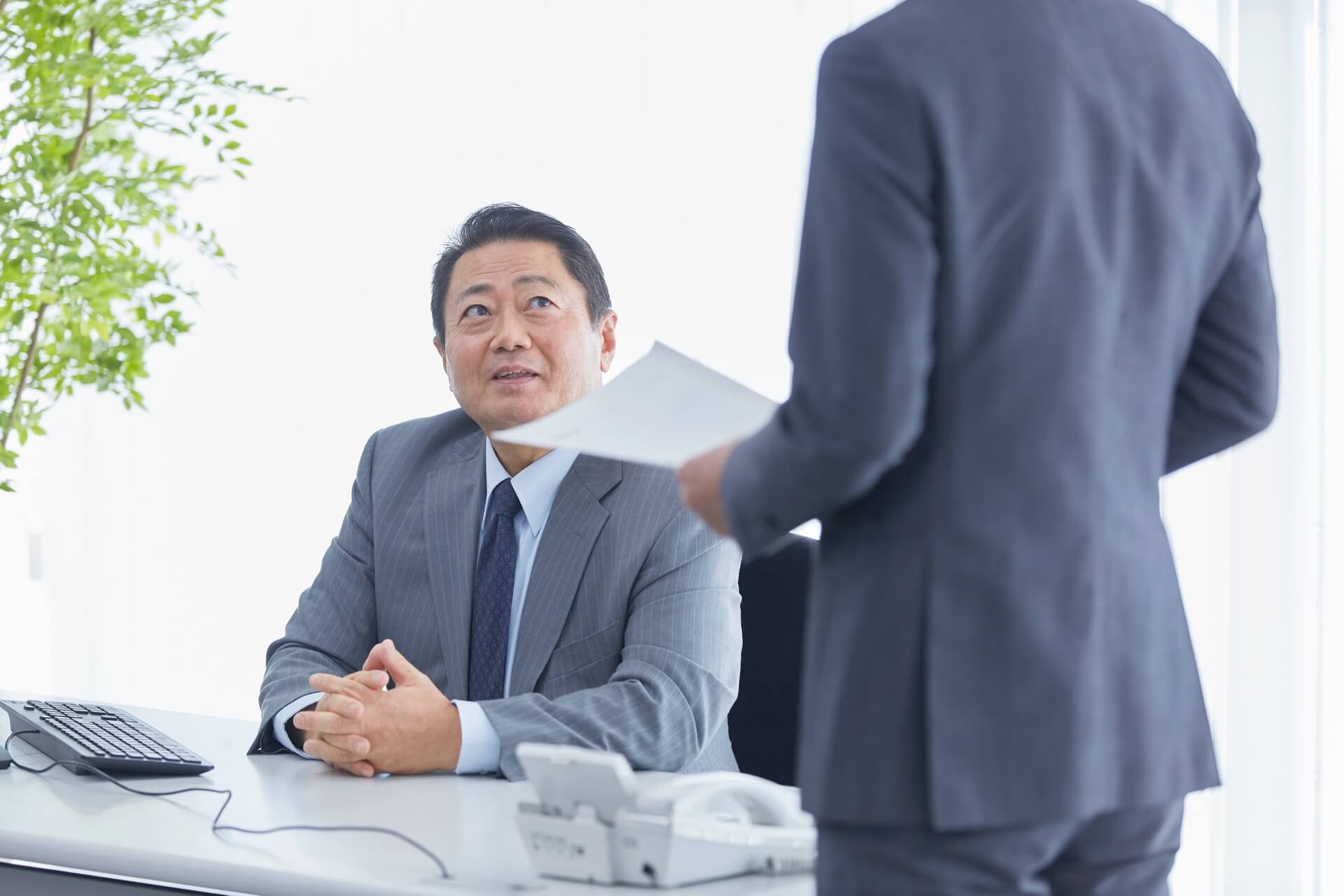 デスクに座る管理職の男性に、書類を持った社員が相談している場面