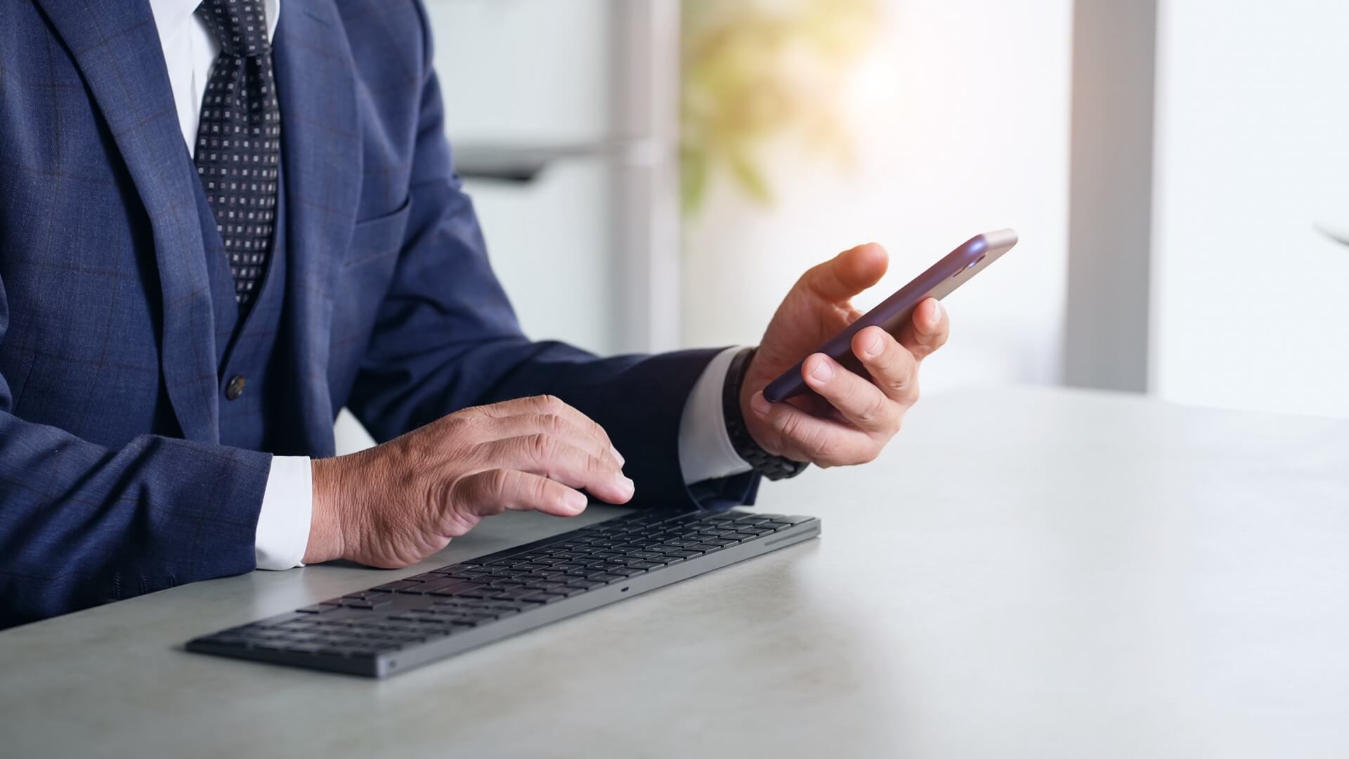 スーツ姿の男性が、ノートパソコンのキーボードに手を置きながらスマートフォンを操作している手元