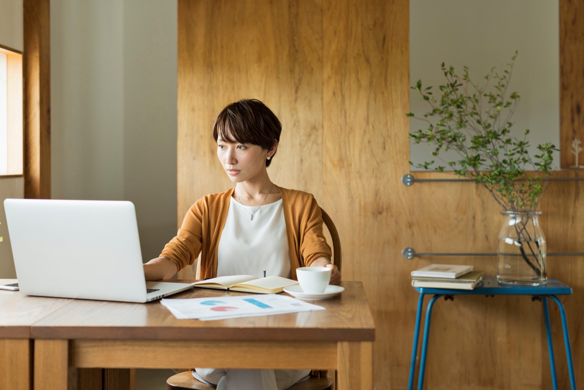 木のぬくもりのある部屋で、ノートパソコンと書類を前に一人で仕事をする女性
